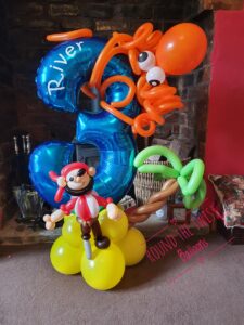 A balloon arrangement featuring a large blue number 3 with the name 'River' inscribed, surrounded by orange balloon octopus, along with a pirate figure made from balloons standing on a yellow base with a palm tree.