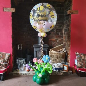A large transparent birthday balloon with bees and flowers, stating 'Happy Birthday Maz', floating above a colourful arrangement of balloon flowers in a green base, set against a rustic fireplace backdrop.