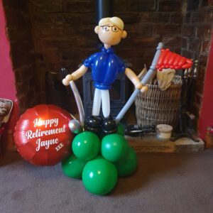A large balloon figure of a man wearing glasses and a blue shirt, standing on a base of green balloons, holding a red flag and tools. Next to him is a round foil balloon that says 'Happy Retirement Jayne' in white letters.