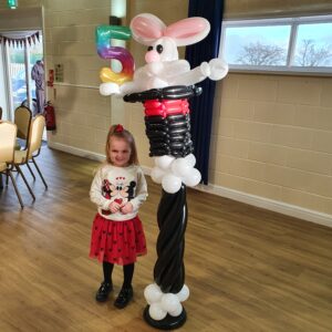 A young girl wearing a Minnie Mouse sweater and a red skirt stands next to a large balloon sculpture of a rabbit emerging from a magic hat, which has a '5' balloon above it, indicating a birthday celebration.