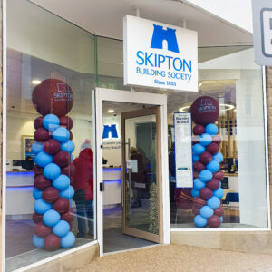 Entrance of Skipton Building Society branch, featuring a prominent sign, colourful balloon columns, and a modern interior visible through the glass door.