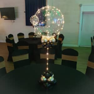 A decorative centrepiece featuring a large transparent globe adorned with fairy lights, atop a cluster of shiny black and silver balloons, set on a black table covered in a fitted tablecloth. The image captures a festive event setting with black chairs having gold sashes.