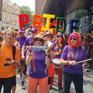 A vibrant group of people celebrating at a pride parade, holding musical instruments and wearing colourful outfits. A large banner made of balloons spells out 'PRIDE' in bright letters.